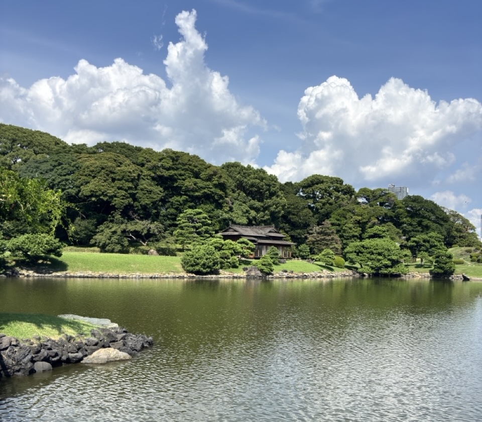 A garden pond with a traditional teahouse on its far bank, office towers visible in the distance.